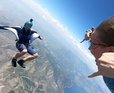 A person skydiving wears a blue jumpsuit and helmet, with arms extended like wings. Another person playfully pretends to squish them from the foreground.