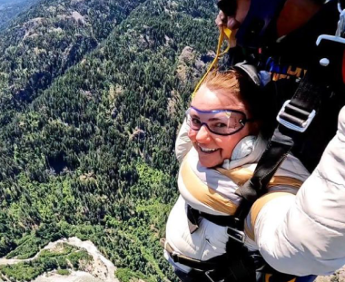 A person is tandem skydiving, smiling widely with goggles on. They're strapped to an instructor against a backdrop of forested hills and clear skies.