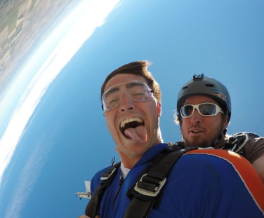 Two people are tandem skydiving; one is ecstatic with their tongue out, the other wears goggles, both against a backdrop of clear blue skies.