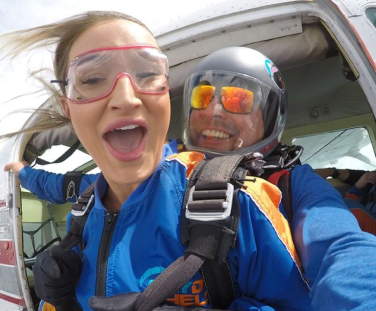 This image shows two excited people skydiving, with the person in the foreground wearing goggles and smiling widely, and the person behind wearing a helmet.