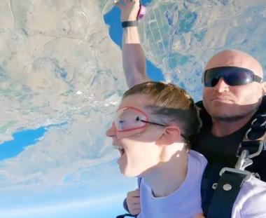 Two people are tandem skydiving, with one person showing an ecstatic expression. The aerial view showcases the earth's surface in the background.