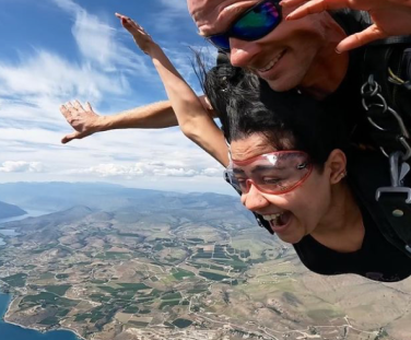 Two people are tandem skydiving above a scenic landscape, featuring green fields, water, and cloudy skies. They seem exhilarated, with expressions of thrill and joy.