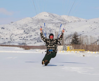 A person is paragliding over a snowy landscape, with a clear blue sky above and snow-covered mountains in the background. They are wearing winter gear.