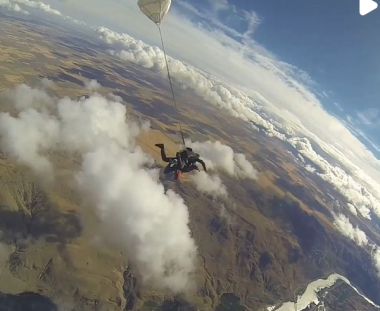 A person is parachuting above a scenic landscape with clouds, rugged terrain below, and a partially deployed parachute visible in the sky.