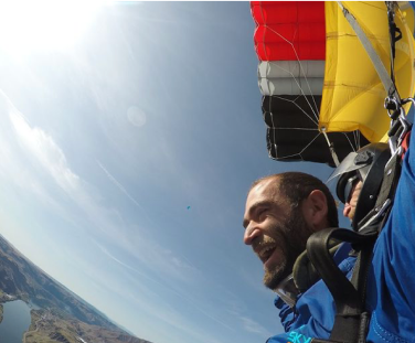 A person is tandem skydiving, smiling with joy. The parachute is visible above, and a landscape with mountains and clear skies is in the background.