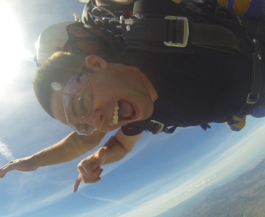 A person is skydiving, captured mid-freefall, with a thrilled expression. They are wearing goggles, a harness, and pointing downward against a clear blue sky background.