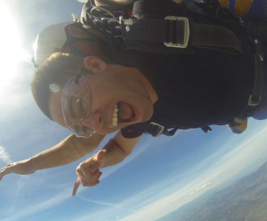 A person is skydiving, captured mid-air with goggles, a harness, and an ecstatic expression. A wide blue sky and distant landscape are visible below.