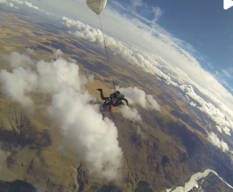 A person is skydiving with a partially deployed parachute above a scenic landscape with mountains, plains, and clouds far below them.