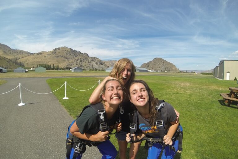 Three people in skydiving gear are posing and smiling for the camera on a grassy field with a hilly landscape and clear skies behind them.