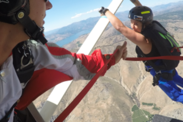 Two people are mid-air engaging in a Skydiving static line progression. One person is hanging onto airplane while the other holds the shoot line.