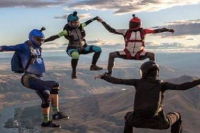 Four skydivers in jumpsuits and helmets are holding hands in formation while free-falling above a landscape with mountains and clouds during sunset.