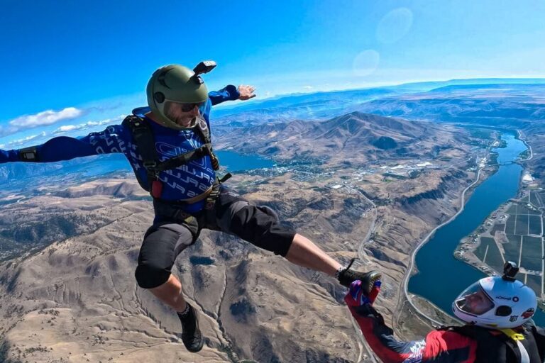 Two people are skydiving above a scenic landscape with a river. One person is in a stable freefall position, while the other captures the moment.