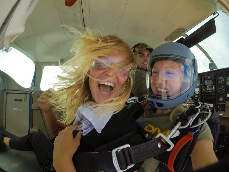 Two people with helmets and goggles are excited inside an aircraft, possibly preparing for a skydive, with equipment strapped on and hair tousled.