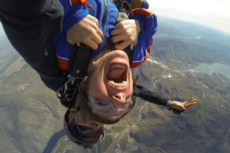 Two people are skydiving in tandem with an open mouthed expression on the foreground person; landscapes are visible below, suggesting high altitude free-fall excitement.